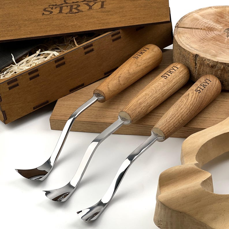 May include: Three metal wood carving tools with curved blades and light brown wooden handles, each branded with the text STRYI. The tools are arranged near a wooden box with the STRYI logo, a wooden block, and a carved wooden piece.
