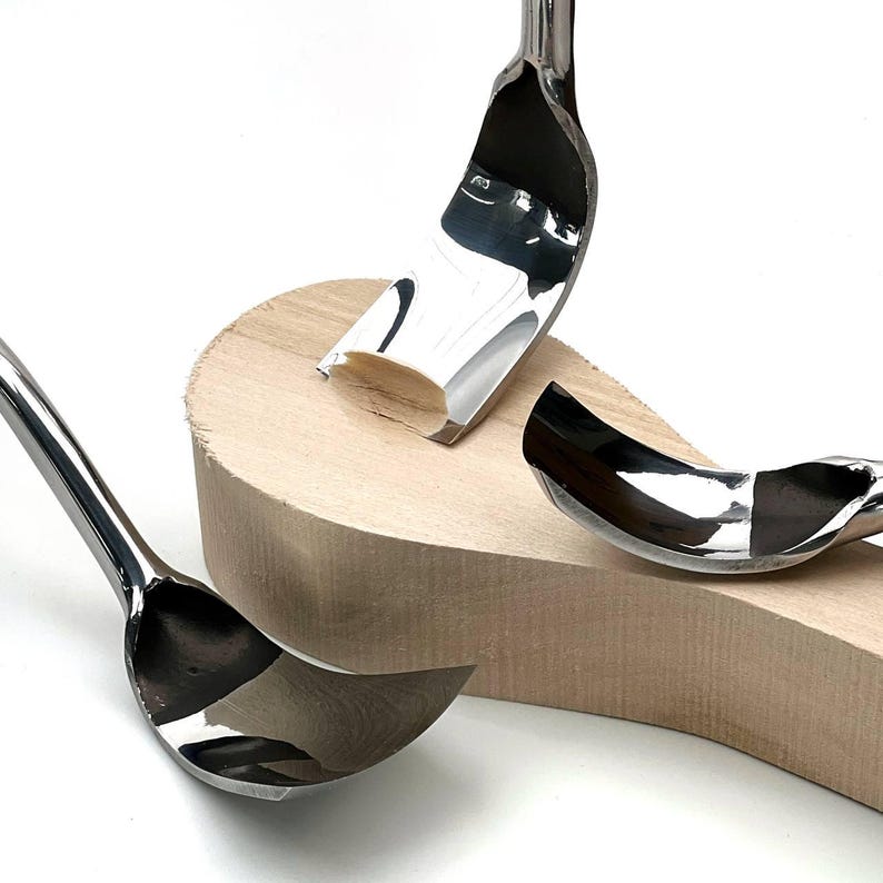 May include: Three polished metal spoon-shaped carving tools with curved, reflective blades resting against a light-coloured, unfinished wooden block. The tools feature smooth, metallic surfaces and distinct, rounded edges designed for shaping wood.