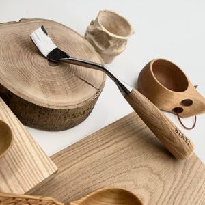 May include: A wood carving tool with a curved, polished metal blade and a light-coloured wooden handle. The handle features the brand name STRYI engraved in black lettering. The tool is positioned near various wooden objects including a round log slice.