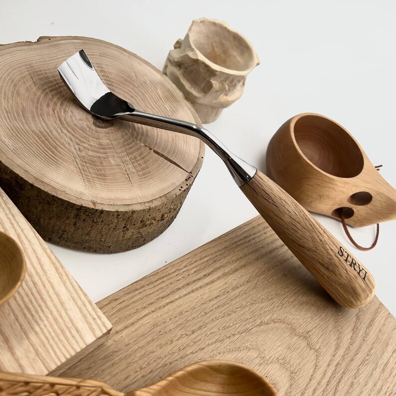 May include: A wood carving tool with a curved, polished metal blade and a light-coloured wooden handle. The handle features the brand name STRYI engraved in black lettering. The tool is positioned near various wooden objects including a round log slice.