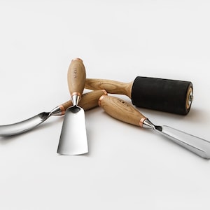 May include: Four wood carving tools with wooden handles and metal blades. The tools include a curved gouge, a flat chisel, a straight chisel, and a wooden roller with a black rubber surface. The handles are all marked with the word "TARIS".