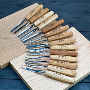 May include: A set of twelve wood carving tools with light-coloured wooden handles, each featuring the brand name STRYI engraved on the side. The tools have various metal blade shapes, including straight, curved, and gouge tips, arranged in a fan pattern.