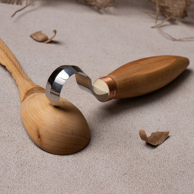 May include: A wooden spoon and a wood carving tool with a metal blade. The tool is resting on top of the spoon.