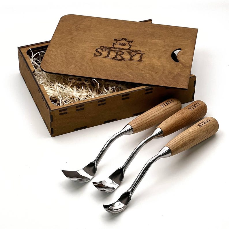 May include: Three metal wood carving tools with light brown wooden handles, each engraved with the word STRYI, arranged in front of an open wooden box containing wood shavings and a lid featuring the STRYI logo and a crown graphic.