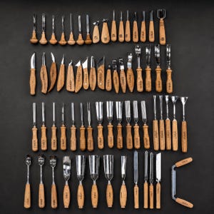 May include: A large collection of wood carving tools with polished metal blades and light brown wooden handles, each stamped with the brand name STRYI. The set includes various gouges, chisels, knives, and a drawknife arranged in rows.