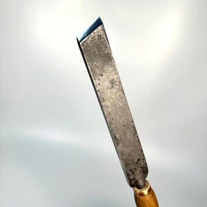 May include: A close-up of a wood handled chisel with a metal blade. The blade is dull and has a patina. The handle is made of light brown wood and has the word "STRYKE" engraved on it.