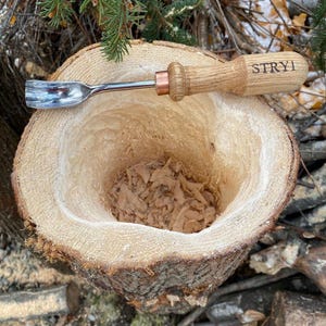 May include: A close-up of a wooden bowl carved from a tree trunk. The bowl is filled with wood shavings. A small, metal carving tool with a wooden handle is resting on the edge of the bowl. The handle is engraved with the word "STRYI".