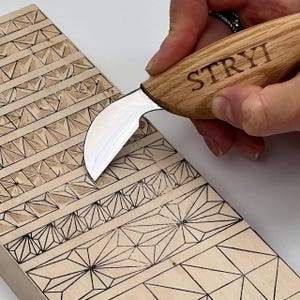 May include: A wood carving knife with a curved blade is held over a wooden block with geometric patterns. The knife handle is engraved with the word "STRYI". The wooden block features various carved designs, including triangles and starburst shapes.
