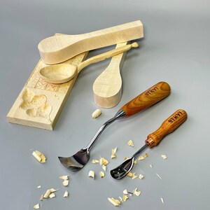 May include: A collection of woodworking tools and wooden spoon blanks. Two spoon carving tools with wooden handles and the text "STRYI" are visible. Several unfinished wooden spoons and a carved wooden block are also present.