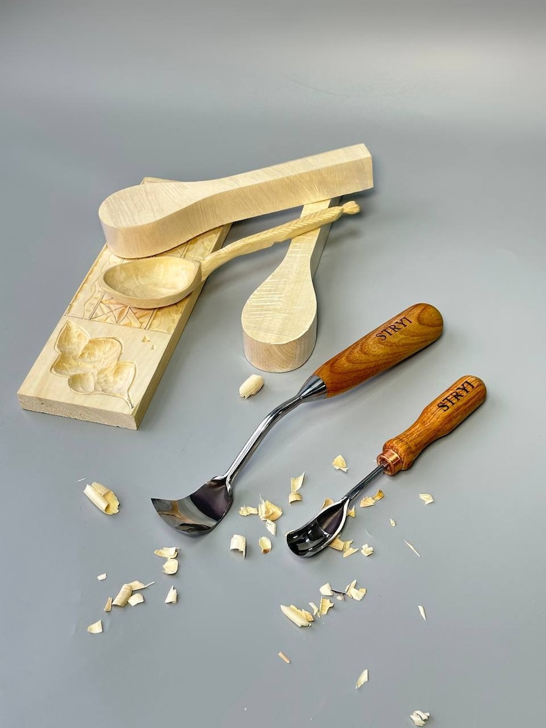 May include: A collection of woodworking tools and wooden spoon blanks. Two spoon carving tools with wooden handles and the text "STRYI" are visible. Several unfinished wooden spoons and a carved wooden block are also present.