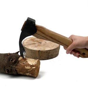 May include: A hand tool with a curved metal blade and a wooden handle, shown striking a piece of rough-cut log. The tool features a dark, matte-finished metal head securely attached to a light brown, grained wooden handle with a visible hole.