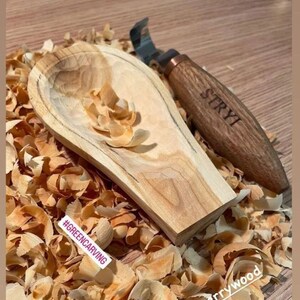 May include: A wooden spoon carved from cherry wood with wood shavings surrounding it. The spoon is unfinished and has a natural wood color. A carving tool with a wooden handle is also in the image. The handle is engraved with the word "STRYI". The image also includes the hashtag "#GREENCARVING".