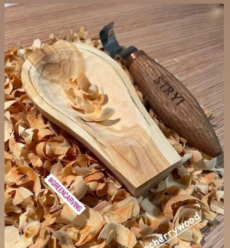 May include: A wooden spoon carved from cherry wood with wood shavings surrounding it. The spoon is unfinished and has a natural wood color. A carving tool with a wooden handle is also in the image. The handle is engraved with the word "STRYI". The image also includes the hashtag "#GREENCARVING".
