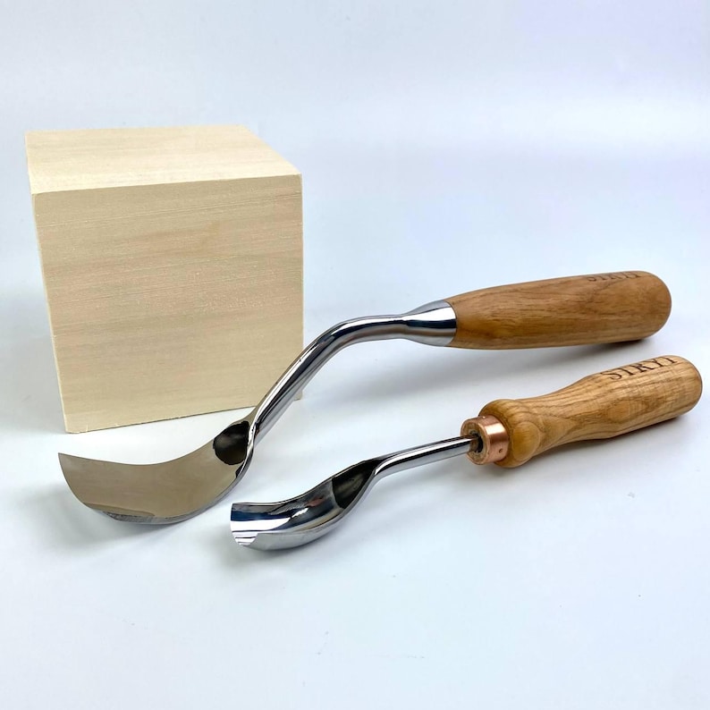 May include: Two wood carving gouges with curved stainless steel blades and wooden handles. The handles are light brown and the blades are shiny. A small, light brown wooden box is in the background.