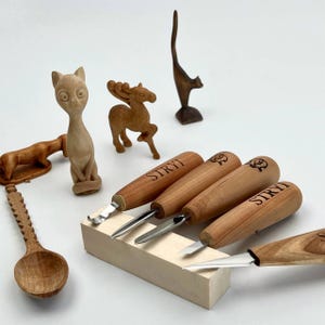 May include: A set of wood carving tools with light brown wooden handles branded with the text STRYI, featuring metal blades of various shapes, alongside several small hand-carved wooden animal figurines and a decorative wooden spoon.