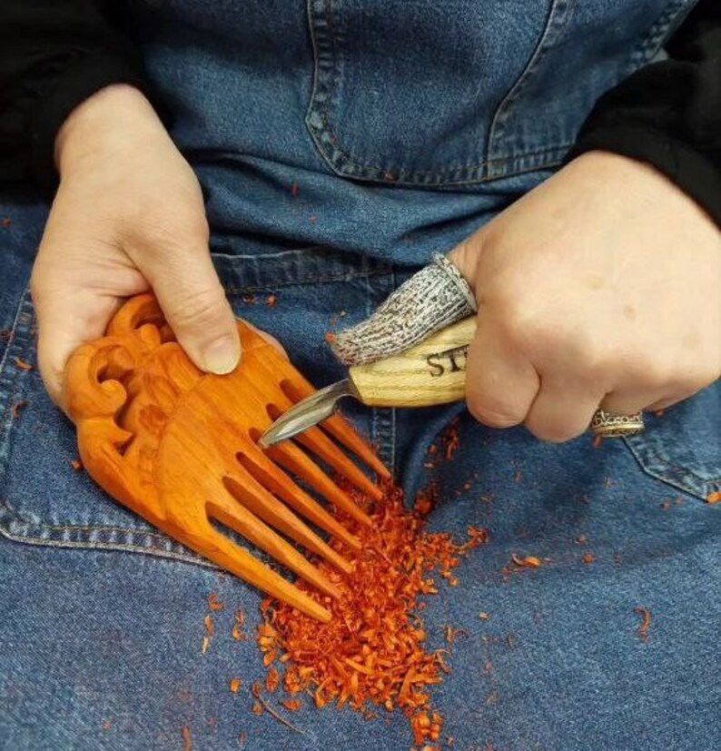 Op de afbeelding: Een persoon is een houten kam aan het snijden met een klein mes. De kam heeft een decoratief ontwerp aan de bovenkant en is gemaakt van roodbruin hout. De persoon draagt een blauwe spijkerbroek en een witte handschoen aan zijn rechterhand.