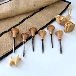 Op de afbeelding: Een set van zeven houtbewerkingsgereedschappen met houten handvatten en metalen bladen. De gereedschappen zijn gerangschikt op een jute oppervlak met een gesneden houten uil en een gesneden houten hond.