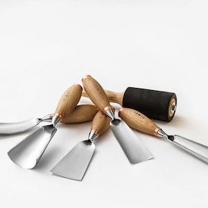 Könnte beinhalten: Ein Satz von fünf Holzschnitzwerkzeugen mit Holzgriffen und Metallklingen. Die Werkzeuge sind auf einer weißen Oberfläche mit einer schwarzen Gummwalze im Hintergrund angeordnet. Die Griffe sind mit "LAWIS" beschriftet.