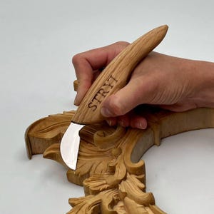 May include: A wood carving knife with a curved blade is held over a carved wooden floral design. The knife handle is made of light-colored wood and has the word "STRYI" printed on it. The carved wood has a natural, light brown color.