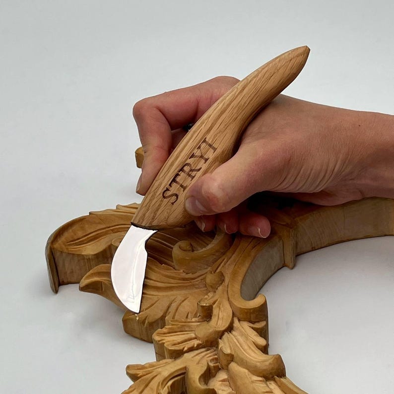 May include: A wood carving knife with a curved blade is held over a carved wooden floral design. The knife handle is made of light-colored wood and has the word "STRYI" printed on it. The carved wood has a natural, light brown color.