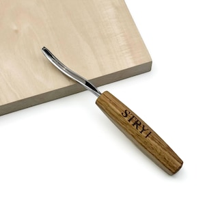Puede incluir: Una gubia para tallar madera con una hoja de metal curva y un mango de madera de color marrón claro. El mango presenta la marca STRYI grabada en letras negras. La herramienta está colocada en diagonal sobre un bloque de madera plano.