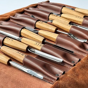 May include: A set of wood carving chisels with light brown wooden handles, each stamped with the text STRY, secured in a brown leather roll-up tool pouch. The metal blades feature various curved and straight edges for detailed woodworking tasks.