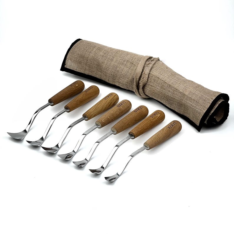 Peut inclure: Un ensemble de sept outils de sculpture sur bois avec des lames en m&eacute;tal poli et des manches en bois brun clair, chacun portant la marque STRYI grav&eacute;e sur le manche. Les outils sont align&eacute;s &agrave; c&ocirc;t&eacute; d'un &eacute;tui de rangement en toile de jute beige.