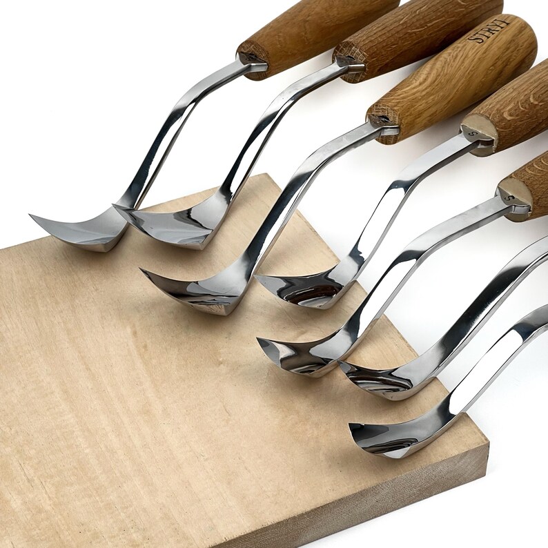 Peut inclure: Un ensemble de cinq outils de sculpture sur bois en m&eacute;tal avec des lames incurv&eacute;es et des manches en bois brun clair. Chaque outil pr&eacute;sente une finition argent&eacute;e polie et un col courb&eacute;. Un manche porte la marque STRYI.