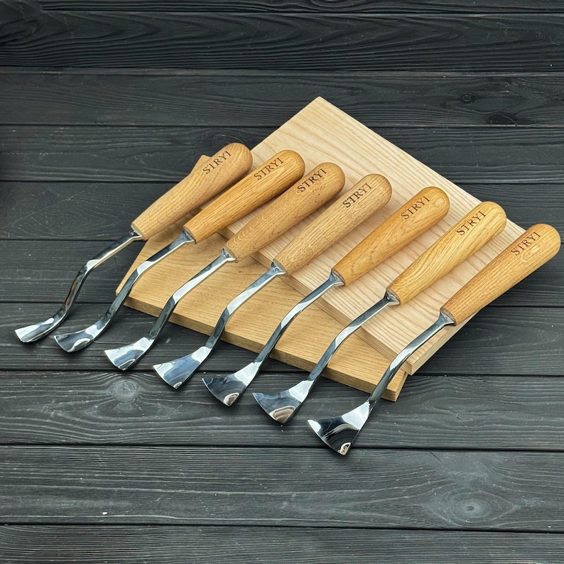 Peut inclure: Un ensemble de six outils de sculpture sur bois avec des manches en bois clair, chacun portant la marque STRYI grav&eacute;e sur le manche. Les outils ont des lames m&eacute;talliques polies et incurv&eacute;es pour le travail du bois, align&eacute;s sur une planche.
