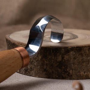 May include: A close-up of a wood carving tool with a curved blade and a wooden handle. The blade is made of polished metal and has a copper ferrule.