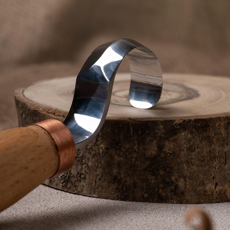 May include: A close-up of a wood carving tool with a curved blade and a wooden handle. The blade is made of polished metal and has a copper ferrule.