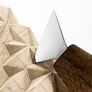 Op de afbeelding: Een metalen houtsnijmes met een driehoekig lemmet en een houten handvat, dat in een lichtgekleurd houten blok snijdt met een geometrisch patroon van uitgesneden driehoekige facetten over het gehele getextureerde oppervlak.