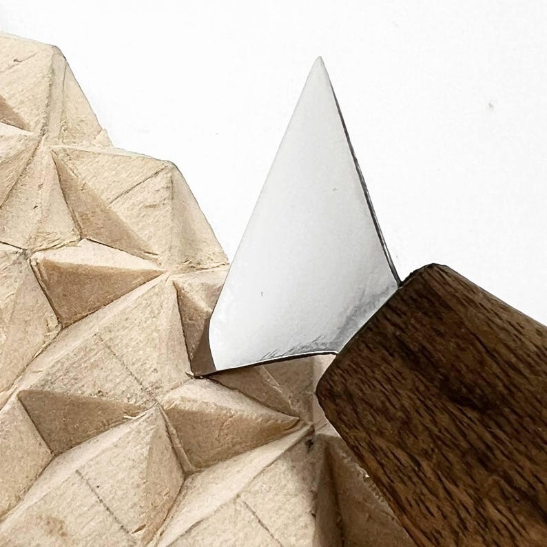 Op de afbeelding: Een metalen houtsnijmes met een driehoekig lemmet en een houten handvat, dat in een lichtgekleurd houten blok snijdt met een geometrisch patroon van uitgesneden driehoekige facetten over het gehele getextureerde oppervlak.