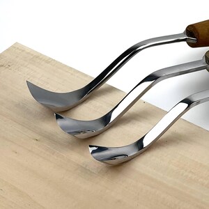 May include: Three metal wood carving tools with curved, polished silver blades and wooden handles. The tools are arranged in a row, showing their distinct hook-shaped tips designed for shaping wood, resting against a light-coloured wooden surface.