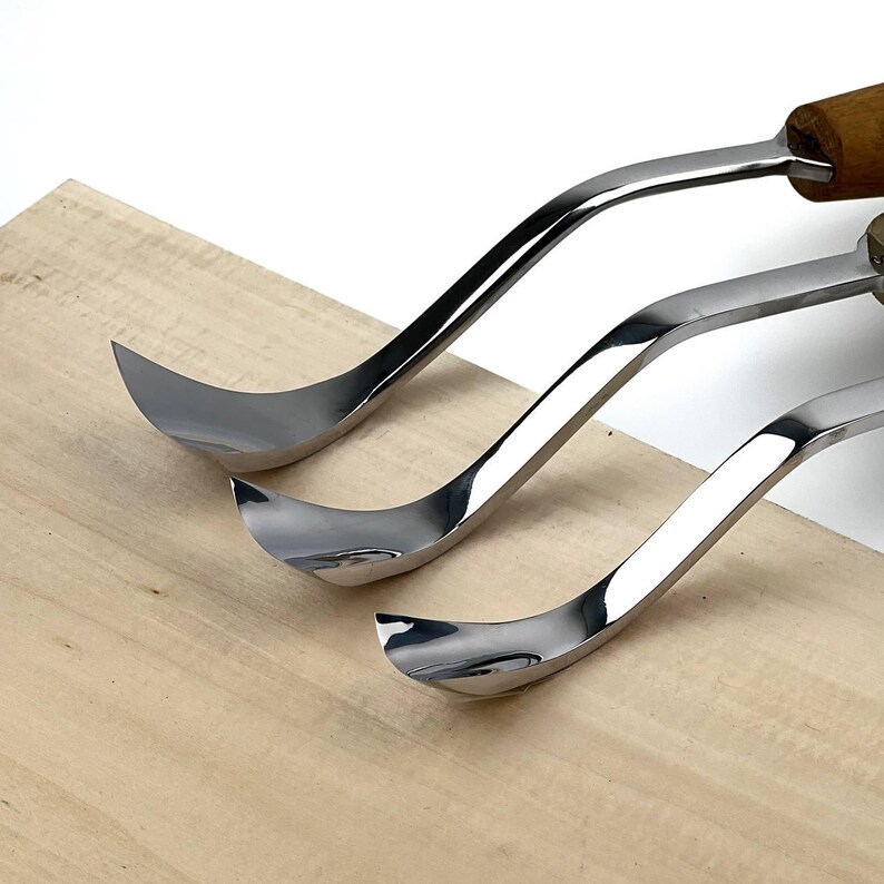 May include: Three metal wood carving tools with curved, polished silver blades and wooden handles. The tools are arranged in a row, showing their distinct hook-shaped tips designed for shaping wood, resting against a light-coloured wooden surface.