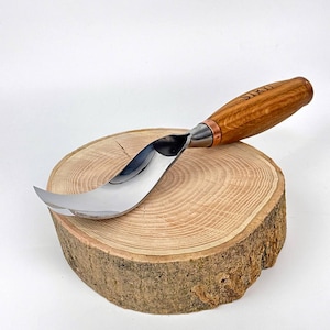 May include: A curved, stainless steel carving knife with a wooden handle. The handle is light brown with the word "SIRU" etched into it. The knife is resting on a round, natural wood slice with visible growth rings.