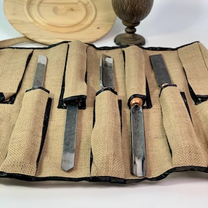 Könnte beinhalten: Ein Satz von sechs Holzschnitzwerkzeugen in einer braunen Leinwandrolle mit schwarzem Besatz. Die Werkzeuge sind aus Metall und haben Holzgriffe.
