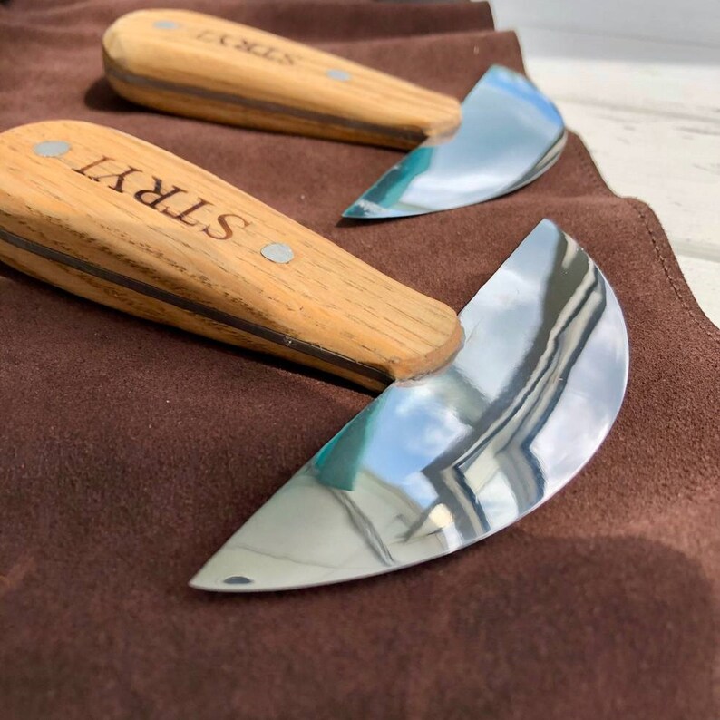 May include: Two leather working tools with wooden handles and curved metal blades. The blades are polished and reflect the surrounding environment. The handles are engraved with the word "STRYI".
