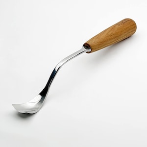 May include: A stainless steel spoon with a curved design and a wooden handle. The handle is light brown and has the word "STRYI" engraved on it. The spoon has a polished, reflective surface.