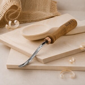 May include: A hook knife with a curved metal blade and a brown wooden handle featuring the engraved text STRYI. The tool is positioned on a light-colored wooden board next to a partially carved wooden spoon and several small wood shavings.