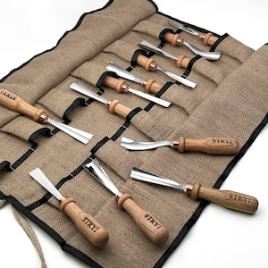 May include: A set of eight wood carving tools with light brown wooden handles, each stamped with the brand name STRYI, featuring polished metal blades of various shapes, organised in a tan burlap roll-up storage case with black trim.