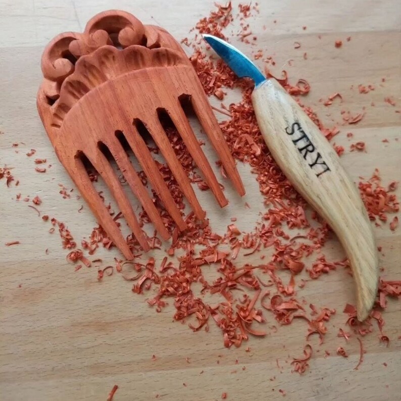 Op de afbeelding: Een handgesneden houten kam met ingewikkelde details en een roodbruine kleur. De kam is omringd door houtsnippers en een klein snijmes met het woord "STRYI" gegraveerd op het handvat.