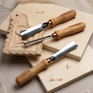 以下が含まれることがあります： 金属製の刃と木製のハンドルが付いた木彫りの道具が3つあります。道具には「STRYI」とラベルが貼られており、木製の表面に置かれています。