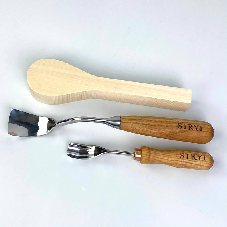 May include: Three wood carving tools are displayed on a white surface. The tools include a wooden spoon blank, a curved gouge with a wooden handle, and a smaller spoon carving tool with a wooden handle. The handles of the tools are engraved with the word "STRYI".