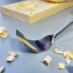 May include: A shiny, curved carving knife with a polished metal handle and blade. Wood shavings are scattered around the tool, suggesting woodworking. A wooden block with carved details is in the background.