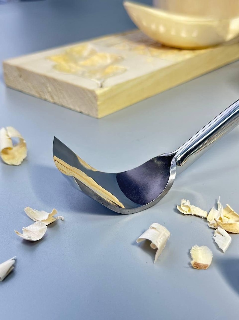 May include: A shiny, curved carving knife with a polished metal handle and blade. Wood shavings are scattered around the tool, suggesting woodworking. A wooden block with carved details is in the background.