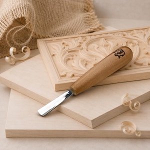 May include: A wood carving chisel with a light brown wooden handle featuring a small black logo, resting on a rectangular wooden board with an intricate carved floral pattern, accompanied by two plain wooden boards and wood shavings.
