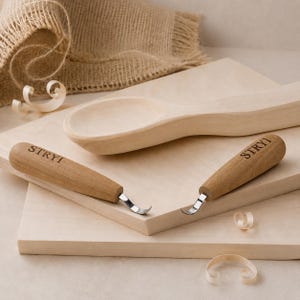 May include: Two wood carving hook knives with light brown wooden handles, each featuring the brand name STRYI engraved in black. The tools have curved metal blades and are positioned on a light-colored wooden board next to a carved wooden spoon.
