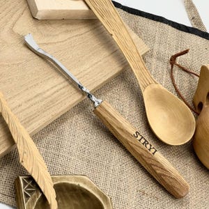 Puede incluir: Una herramienta de tallado de madera con una cuchilla metálica curva y un mango de madera. El mango está grabado con la palabra "STRYI". También se ven otras herramientas de tallado de madera y una cuchara de madera.