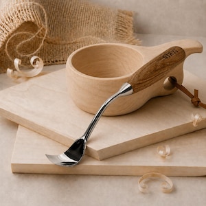 May include: A silver metal hook knife with a wooden handle featuring the text STRYI, positioned next to a light-colored wooden cup with a handle and a leather cord. The tool has a curved blade and a smooth, polished finish on the metal surface.
